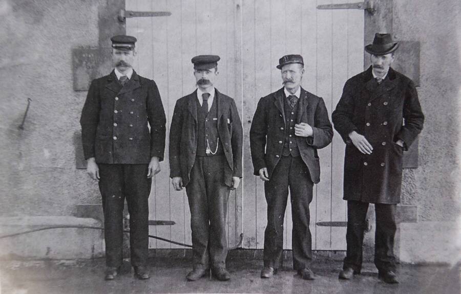 Black and white photograph. Four men in a row; the first three are the Flannan Isle lightkeepers, from left to right James Ducat, Thomas Marshall and Donald McArthur. The last man is the Superintendent Robert Muirhead