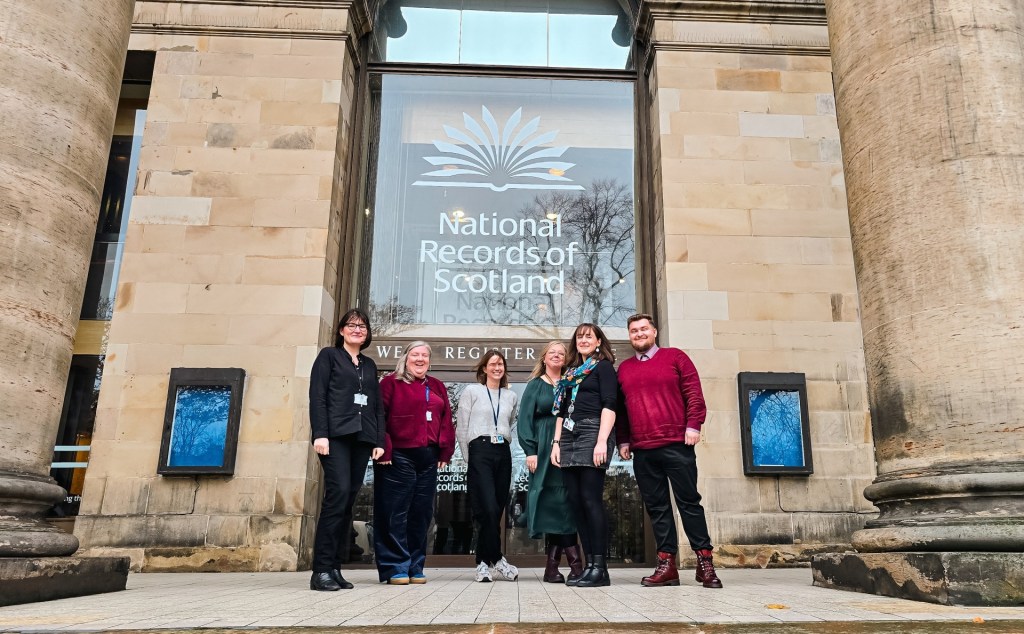 Six members of the digital archiving branch standing outside the West Register House Building in Edinburgh with a large glass window behind them. They're framed by sandstone columns. 