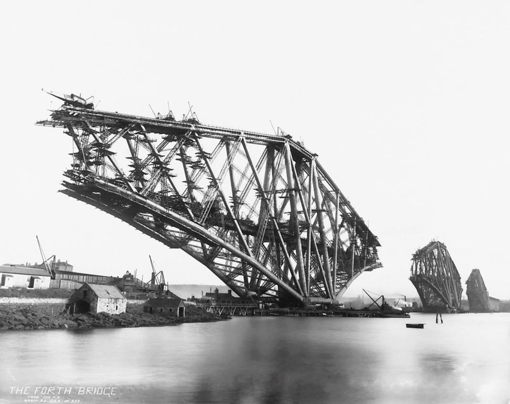 The Forth Bridge under construction, showcasing the steel structure and cranes, with the river and nearby buildings visible.